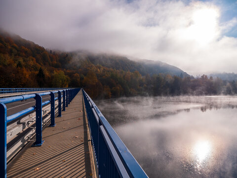 Pedestrian walkway next to road on the Ružín bridge near Košické Hámre, Slovakia. Autumn morning fog over reservoir with forested hills and sun reflection.