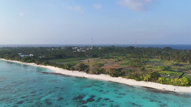 Aerial view of Thoddoo Island's white sandy beach meeting turquoise water, lush green vegetation, and scattered buildings, Thoddoo, Alif Alif Atoll, Maldives.