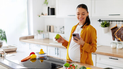 Happy young pregnant woman using her smartphone and having snack, eating apple standing in well-lit kitchen interior