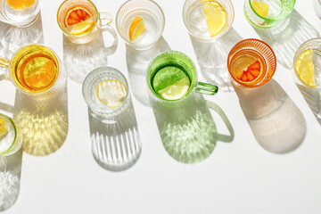 Summer still life with colored glass cups of infused water with slices of fruit on white table. Glittering different glasses at sunlight. Long harsh shadow. Summer vacation concept, top view