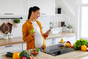 Smiling pregnant woman standing in modern kitchen holding apple in one hand and using cellphone...
