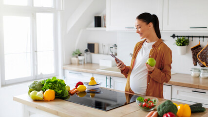 Smiling pregnant woman standing in modern kitchen holding apple in one hand and using cellphone...