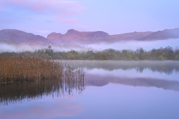 Fototapeta premium Misty Elterwater lake on an autumn morning with calm mountain reflections, Lake District, UK.