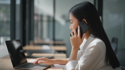 A businesswoman making a phone call while using a laptop in a modern office setting. - Powered by Adobe