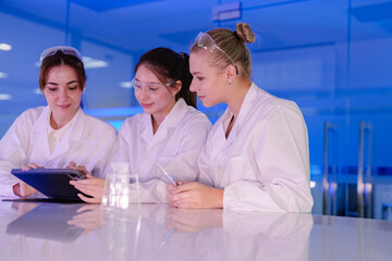 A team of three young female scientists smiles while working together in a futuristic, blue-lit lab. They are looking at a tablet, sharing ideas and discussing their research.