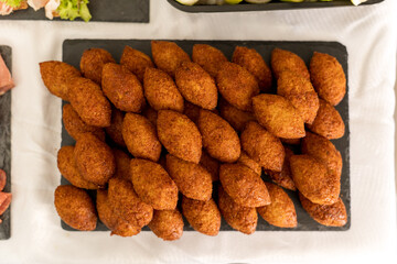 A large platter holds a stack of golden-brown fried kibbeh, perfectly shaped and arranged. The festive atmosphere is enhanced by soft lighting and nearby food offerings.