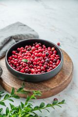 Fresh cranberries in a dark bowl on a wooden board on a light marble background with green branch