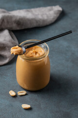 Creamy peanut butter in a jar on a dark background with roasted nuts spoon and napkin.