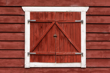A charming wooden window with crossed shutters is set against the vibrant red walls of a barn. The scene captures the essence of rural life and tranquility in the countryside.