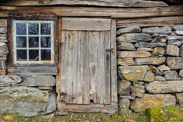 A weathered wooden door stands beside a stone wall of a cottage. The window is framed by old wood, revealing the peaceful countryside on a clear autumn day.