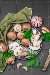 Fresh brown mushrooms with garlic, herbs and rustic knife on black tile kitchen surface