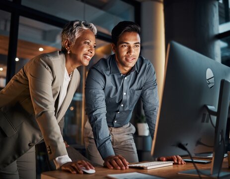 Mature female executive mentoring a younger male colleague at a computer, both smiling and collaborating on a project in a modern office at night. - Powered by Adobe