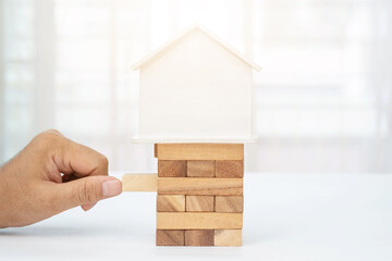 A man's hand holds a wooden block with a house placed on a pile of wood.