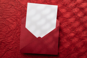 One red envelope, white card textured dark red background with heart-shaped shadows. Valentine's Day, Mother's Day, Women's Day, Wedding, Anniversary, or Birthday.  Flat lay with copy space.