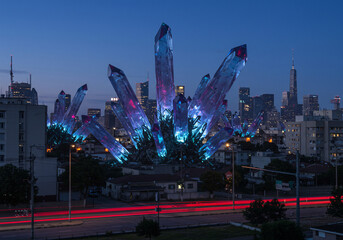 Large glowing blue and purple crystals emerge from a city park at night, with blurred car lights and a cityscape in the background.