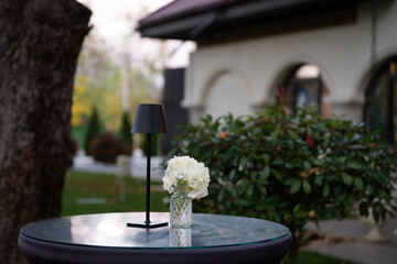 Charming floral arrangement in a small glass vase placed next to a modern black lamp in the outdoor courtyard of a restaurant. The elegant composition combines natural beauty and warm lighting, creati