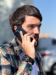 Close-up portrait of a thoughtful young man talking on a smartphone outdoors on a sunny day, wearing a wristwatch.