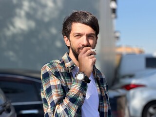 Portrait of a young man smoking a cigarette outdoors on a sunny day, with an urban background....