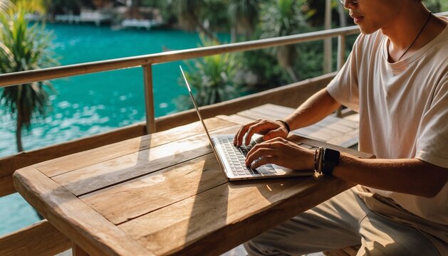 Digital nomad working on a laptop at a wooden table on a balcony with a stunning tropical blue water view. - Powered by Adobe