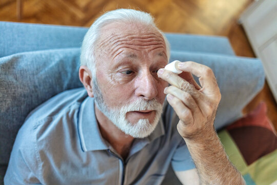 An elderly retired man is at home, sitting on the sofa and instilling medicine in his eye for inflammation and fatigue. A Senior man with cataract or conjunctivitis is using an eye drops at home.