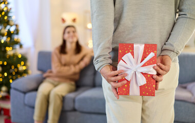 Christmas couple sharing gift at cozy home. A man hides a present behind back, planning a holiday...