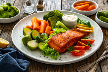 Fried salmon steak with boiled broccoli, brussels sprouts and carrots and mayonnaise on wooden table	