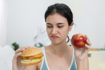 Women looking conflicted while choosing between healthy red apple and chicken burger, expressing...