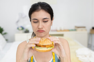 Unhappy women holding fried chicken burger while wearing measuring tape, showing frustration with...