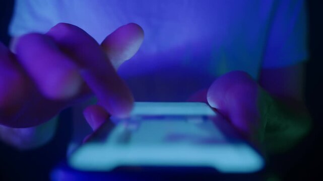 Close-up of male hands using a smartphone, scrolling through social media content on a glowing screen in low light with blue neon illumination. Screen addiction and digital dependence concept