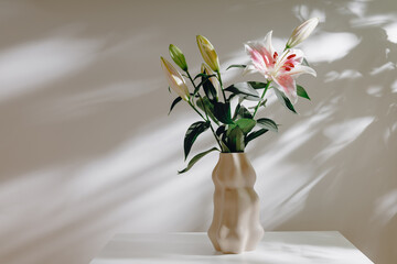 Aesthetic vase with lily flowers bouquet on the white table with beautiful shadows. Relaxing harmonious still life composition, minimal decor for home interior or poster