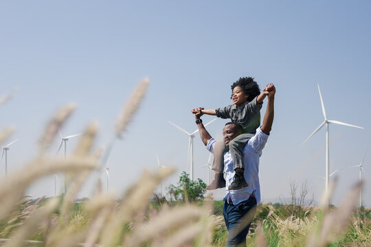 Happy father carrying daughter on shoulders in wind farm. Family bonding, outdoor fun, and clean energy concept. Wind turbines in background symbolize sustainable future and green power.