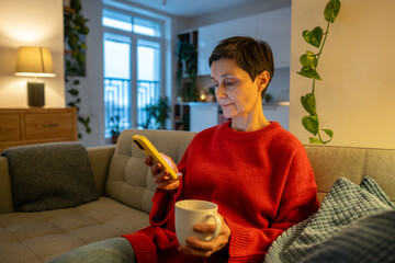 Senior woman sitting comfortably on sofa at home at evening with mug of drink and scrolling social media on smartphone. Mature female relaxing on couch with phone and coffee or tea, typing message.