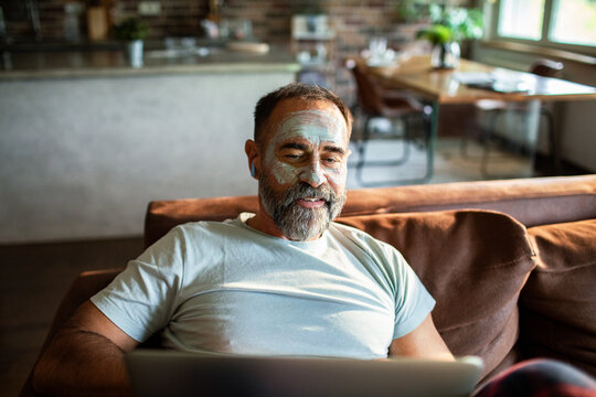 Middle aged man with a face mask using a laptop on the couch at home - Powered by Adobe