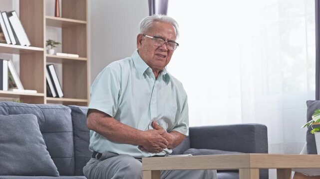 Asian Elderly man clutching abdomen seated living room sofa discomfort senior experiencing stomach pain and concern while resting home elderly man holding abdomen pain seated living room sofa with