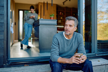 Mature man sitting outside his modern home, holding a cup of coffee and looking away, while a woman uses a phone indoors