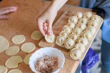 Close-up of woman’s hands placing dumplings in order on wooden board beside bowl of filling. Concept of precision, care, and traditional home cooking with copy space.