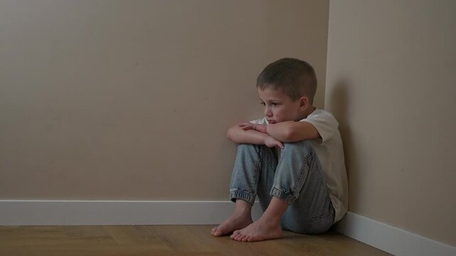 Boy sitting alone in quiet room corner with arms around legs. Child keeps head low, avoiding eye contact. Emotion appears in child still posture. Thinking boy remains silent in space filled emotion.