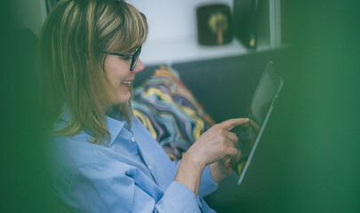 Woman working remotely. Uses digital tablet on sofa. Comfortable work from home. Relaxed workspace