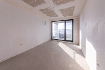Interior of an empty room under construction in a new apartment. Unfinished space with raw concrete walls, floor, and a large window. Renovation and real estate concept.