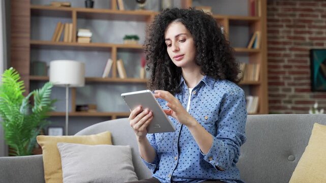Young woman using digital tablet sitting on sofa in living room at home. Beautiful female student browses social media, surfs web, chats online, writes or reads messages holding a device in her hands
