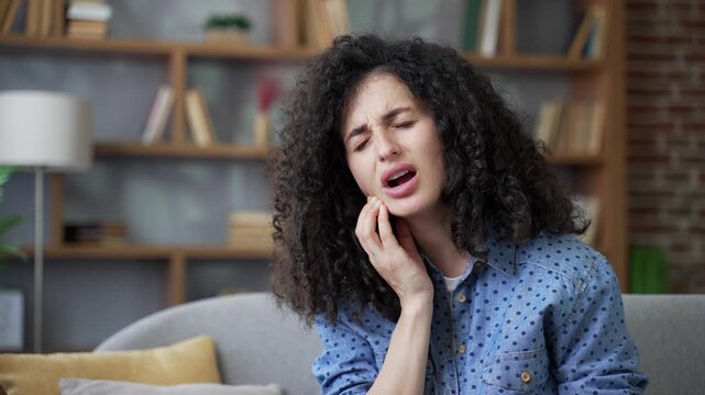 Unwell young woman suffering from toothache sitting on sofa in living room at home. Upset female holds her hand on her cheek, massaging sore spot. She feels sharp pain, needs to see a dentist. Close u