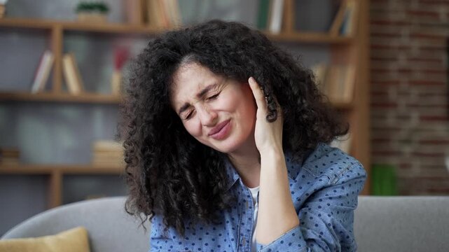 Unhealthy young woman suffers from ear pain sitting on sofa at home. Unwell female touches her ear with fingers. She has furuncle, shingles, eczema, ear diseases or otitis media. Earache. Close up