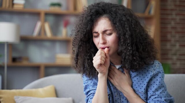 Sick young woman coughing while sitting on sofa in living room at home. Unhealthy adult female feels bad, holds her hand to chest. Unwell lady has a cold, virus, flu, bronchitis or pneumonia. Close up