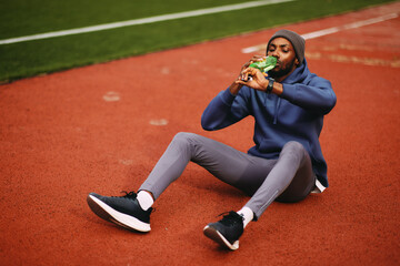 Male athlete sits down on a running track to hydrate, drinking water from a green bottle during a focused recovery break after an outdoor workout.
