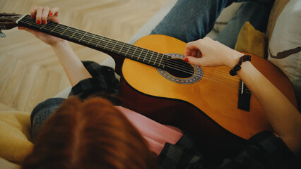 Young woman playing acoustic guitar while sitting comfortably on a sofa in a cozy living room...