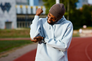 Close-up of an African American male athlete clenching his teeth in intense pain while holding his injured elbow or shoulder during a recovery break on a running track