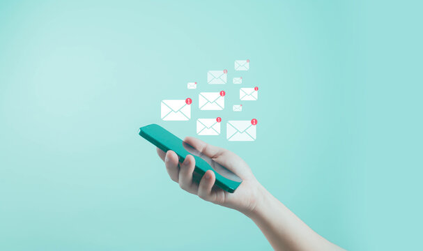 Hand holding smartphone with multiple email notification icons appearing on turquoise background, representing online communication, digital marketing, messaging, social connection, modern technology