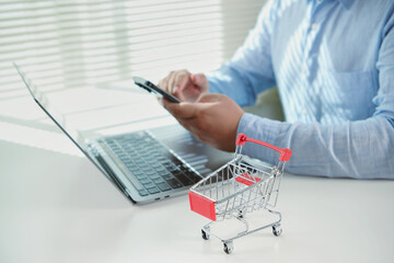 Online shopping concept with businessman using laptop and smartphone alongside shopping cart symbolizing e-commerce digital marketplace mobile payment retail technology and consumer convenience