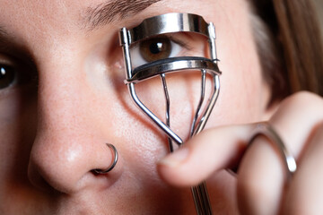 Close-up of a young Caucasian woman using a metal eyelash curler. She has brown hair and a nose ring. The focus is on her eye and the curler.