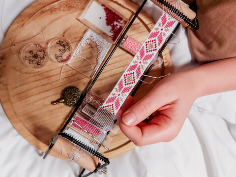 Weaving a bracelet on a loom with a pattern of national Ukrainian motifs in pink tones, creating jewelry from beads as a hobby